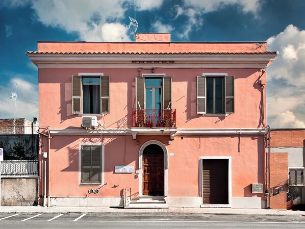 A two-story building with a pink facade, a wooden door, and a balcony with red railings.