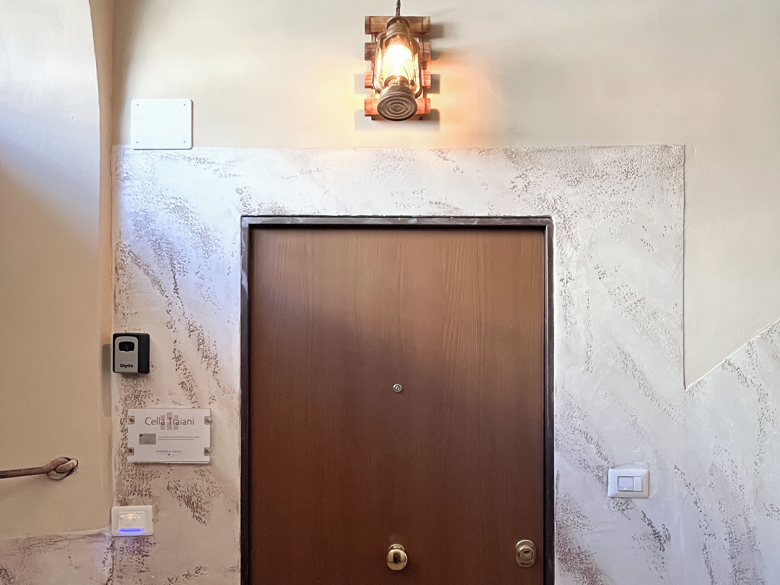 Una puerta de madera con un ojo de cerradura y una cerradura, un aplique en la pared encima y un enchufe a la izquierda.