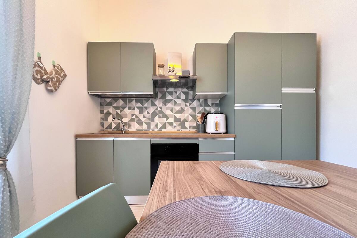 A compact kitchen with grey cabinets, a patterned backsplash, and a wooden dining table with a placemat.