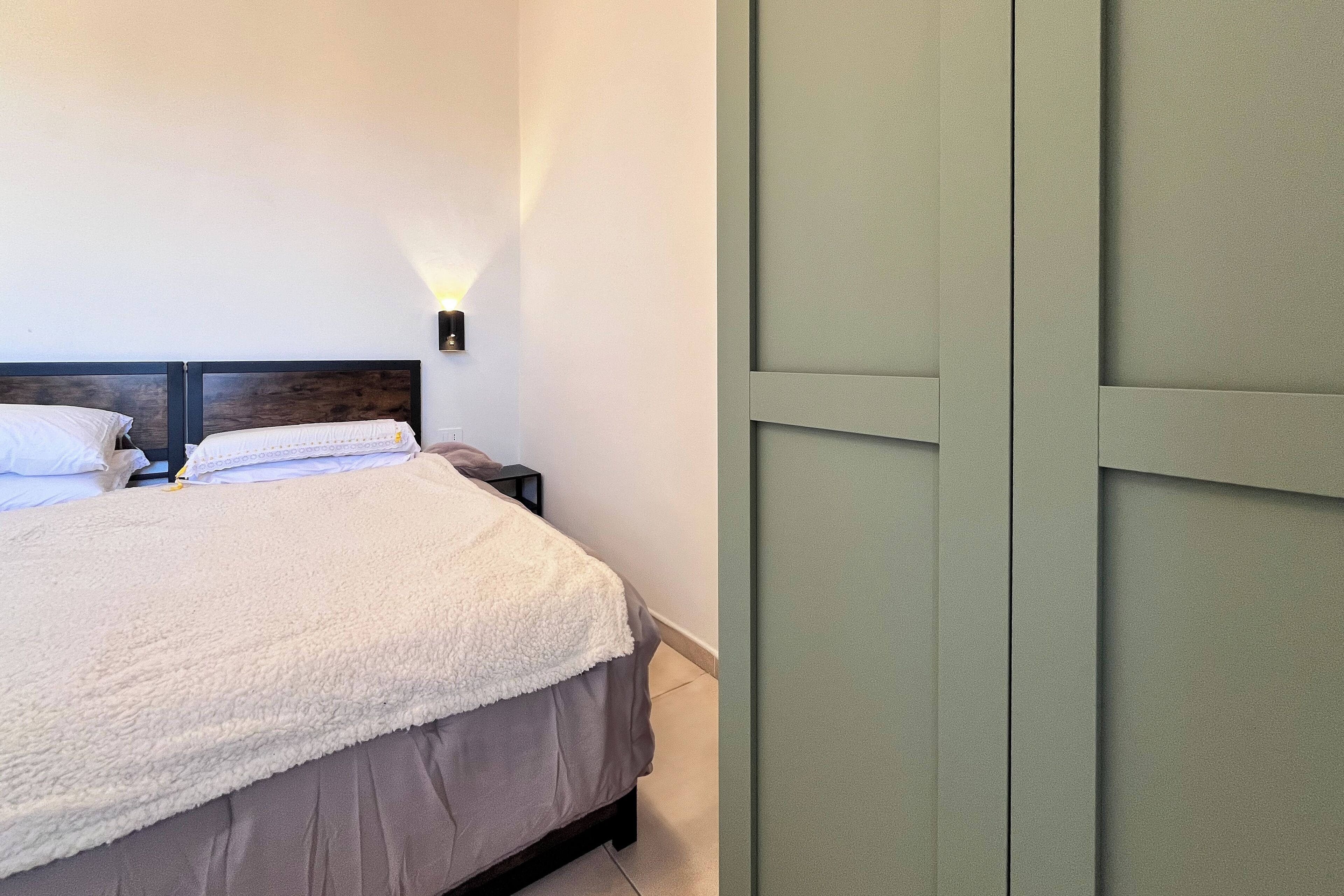 Uma cama bem arrumada com um cobertor branco e cabeceira de madeira, uma mesa de cabeceira com abajur e uma porta verde.