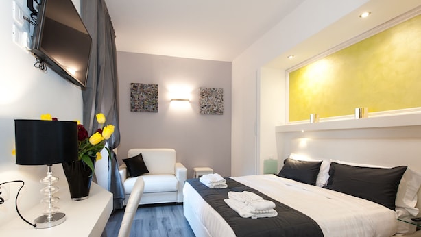 A hotel room with a bed, a chair, a desk with a lamp, and artwork on the walls.