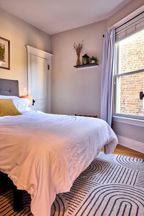 Ein Schlafzimmer mit einem großen Bett, einem Nachttisch, einem Fenster mit Vorhängen, einer Backsteinwand und einem Regal mit Dekoration.