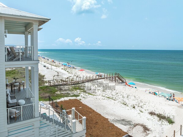 A beach house with a deck, outdoor seating, and a view of the beach and ocean.