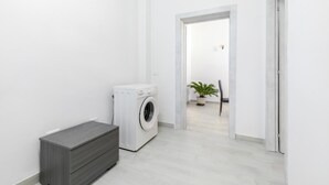 Een wasserette met een wasmachine, een grijs kastje en een bloempot op de achtergrond.