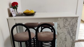 Een keukenhoek met een wit aanrecht, drie zwarte barkrukken, een schaal met fruit en een vaas met rode bloemen.