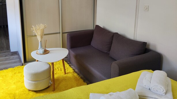 Een hotelkamer met een bank, een klein rond tafeltje met een vaas, een geel tapijt en handdoeken op een bed.