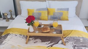 Een net opgemaakt bed met een deken met een geel-wit patroon, een dienblad met croissants en sinaasappelsap, en een rode roos op de nachtkastje.