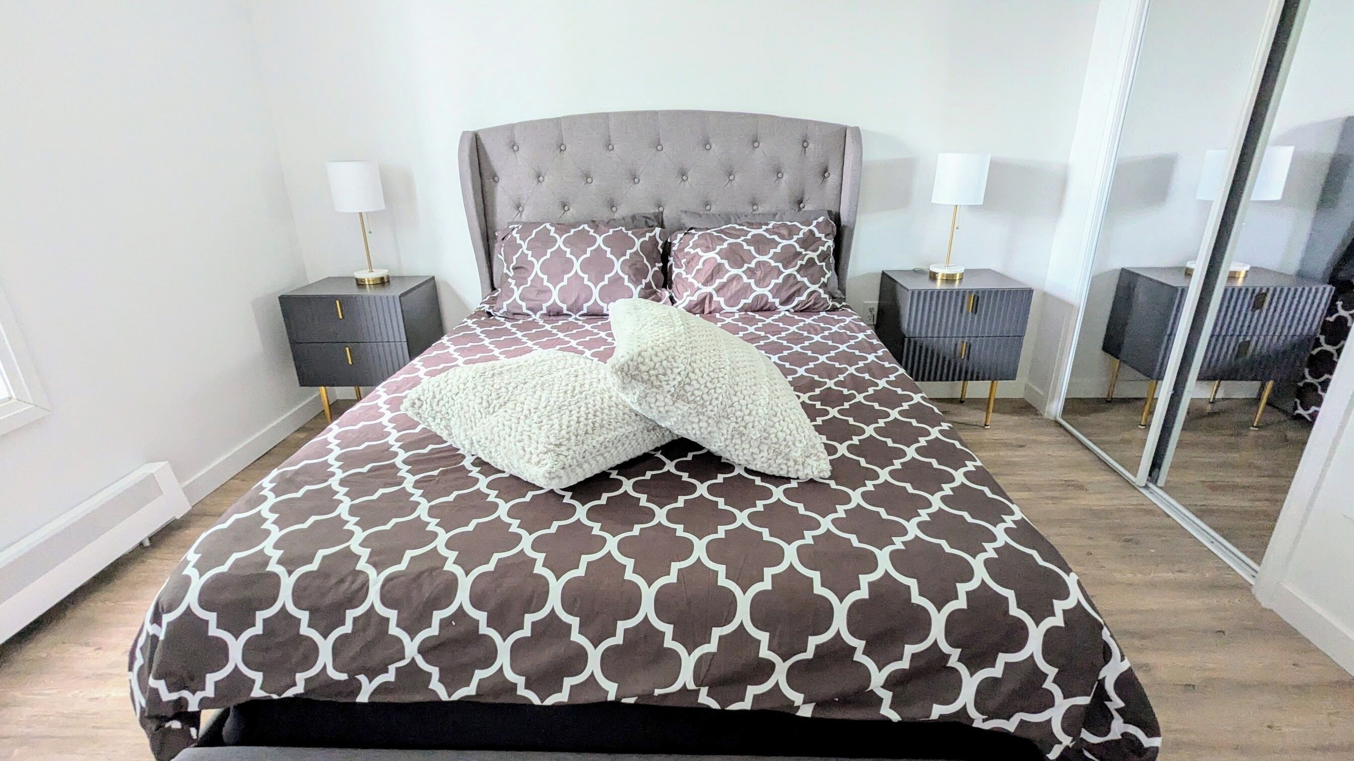 Una cama bien hecha con una colcha estampada, dos almohadas decorativas y un cabecero acolchado. Hay dos mesitas de noche con lámparas y un espejo en la pared.