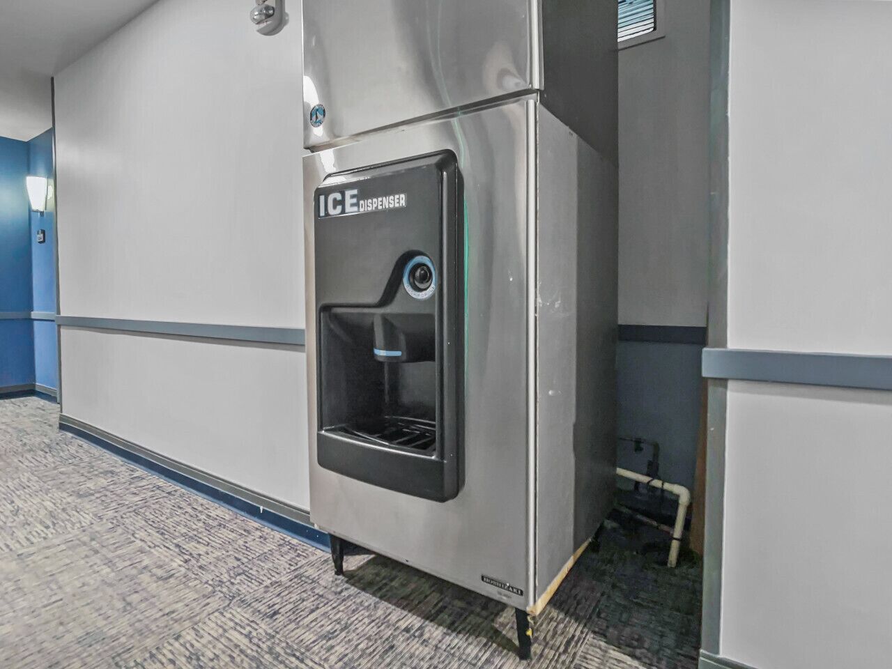 A metallic ice dispenser in a hallway with a grey floor and walls.