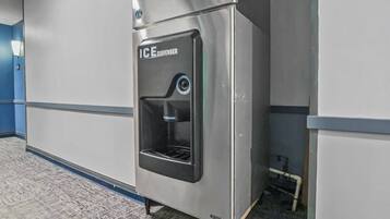 A metallic ice dispenser in a hallway with a grey floor and walls.