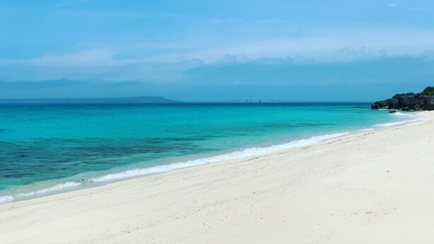 A beach with clear turquoise water, a sandy shore, and distant rocky outcrops. - Miyakojima White Shisa (Miyakojima)