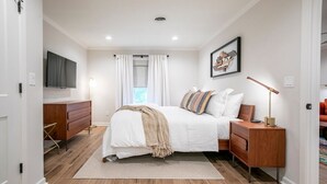 Een slaapkamer met een bed, nachtkastjes, een dressoir, een televisie en een raam met gordijnen.