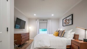 A bedroom with a bed, bedside tables, a TV, a window with curtains, and a painting on the wall.