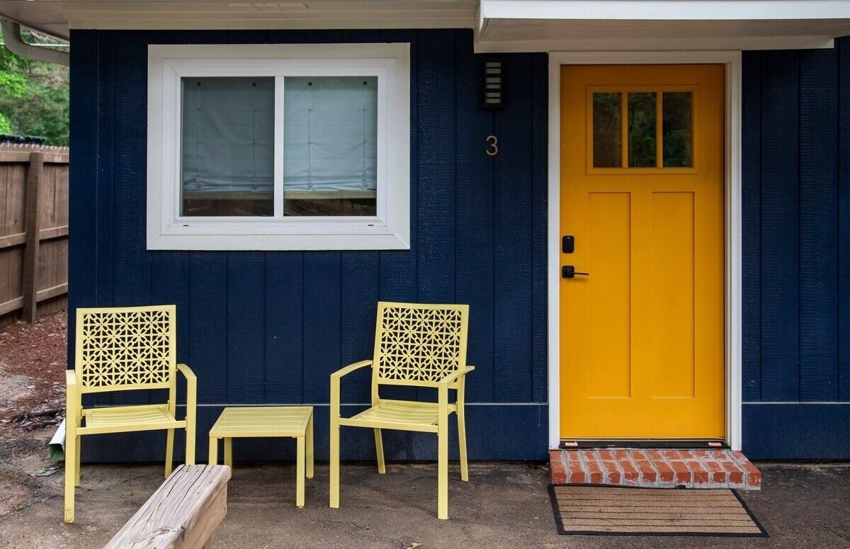 A house with a yellow door, a window with blinds, and two yellow chairs and a small table outside.