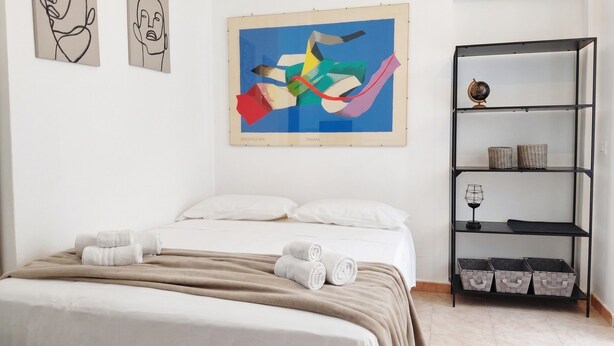 A bedroom with a bed, pillows, a blanket, a shelf with baskets, and a colorful abstract painting. - Casa Olmedina - Studio Apartment, Sleeps 2 (Olmedo)