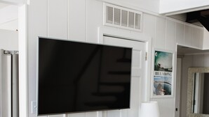 A modern interior with a large flat-screen TV mounted on a white wall, a wooden console table with storage baskets, and a dark wooden floor. - A-Frame - 2 minute walk to Beach - Pet Friendly - HUGE front and back porch (Carolina Beach)