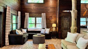 A cozy cabin-style room with a wooden interior, a large window with curtains, a round wicker ottoman, and a wooden coffee table. - Family Fun! Diving Board/Water Slide/Boat Bar/ Beach/Jump Platforms/Green Lights (Eclectic)