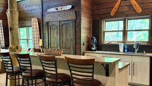 A rustic kitchen with wooden walls, a bar area with stools, and a large window with a view of trees. - Family Fun! Diving Board/Water Slide/Boat Bar/ Beach/Jump Platforms/Green Lights (Eclectic)