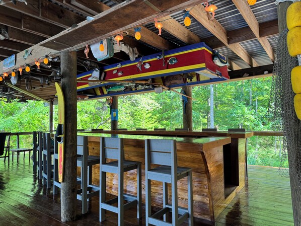 A rustic wooden structure with a wooden deck, a covered seating area with metal chairs, and a view of lush greenery outside. - Family Fun! Diving Board/Water Slide/Boat Bar/ Beach/Jump Platforms/Green Lights (Eclectic)