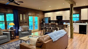 A cozy living room with a brown leather sofa, a coffee table, and a TV. There is a kitchen area with wooden cabinets and a dining area with bar stools. - Family Fun! Diving Board/Water Slide/Boat Bar/ Beach/Jump Platforms/Green Lights (Eclectic)