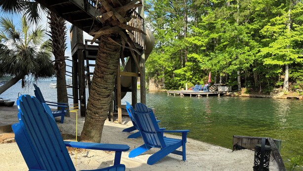 A beachside area with blue Adirondack chairs, a palm tree, and a dock extending into a lake surrounded by greenery. - Family Fun! Diving Board/Water Slide/Boat Bar/ Beach/Jump Platforms/Green Lights (Eclectic)