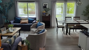 Un vaste espace de vie comprenant un canapé, des fauteuils, une table à manger et un coin cuisine. De grandes fenêtres laissent entrer la lumière naturelle et offrent une vue sur la verdure environnante.