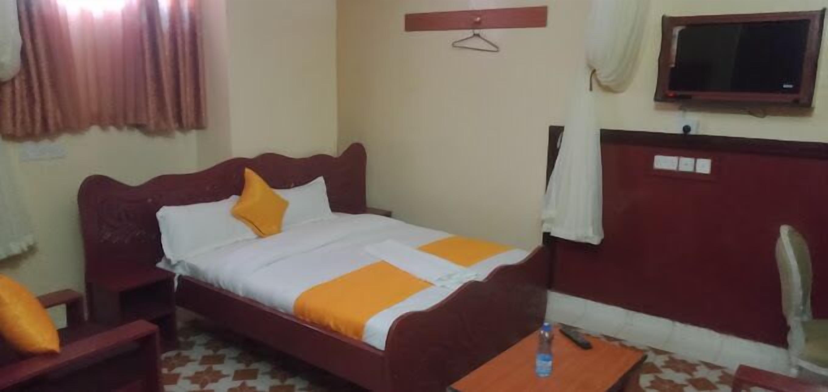 A single bed with a wooden headboard, a bedside table, and a chair. There is a television mounted on the wall and a window with curtains.