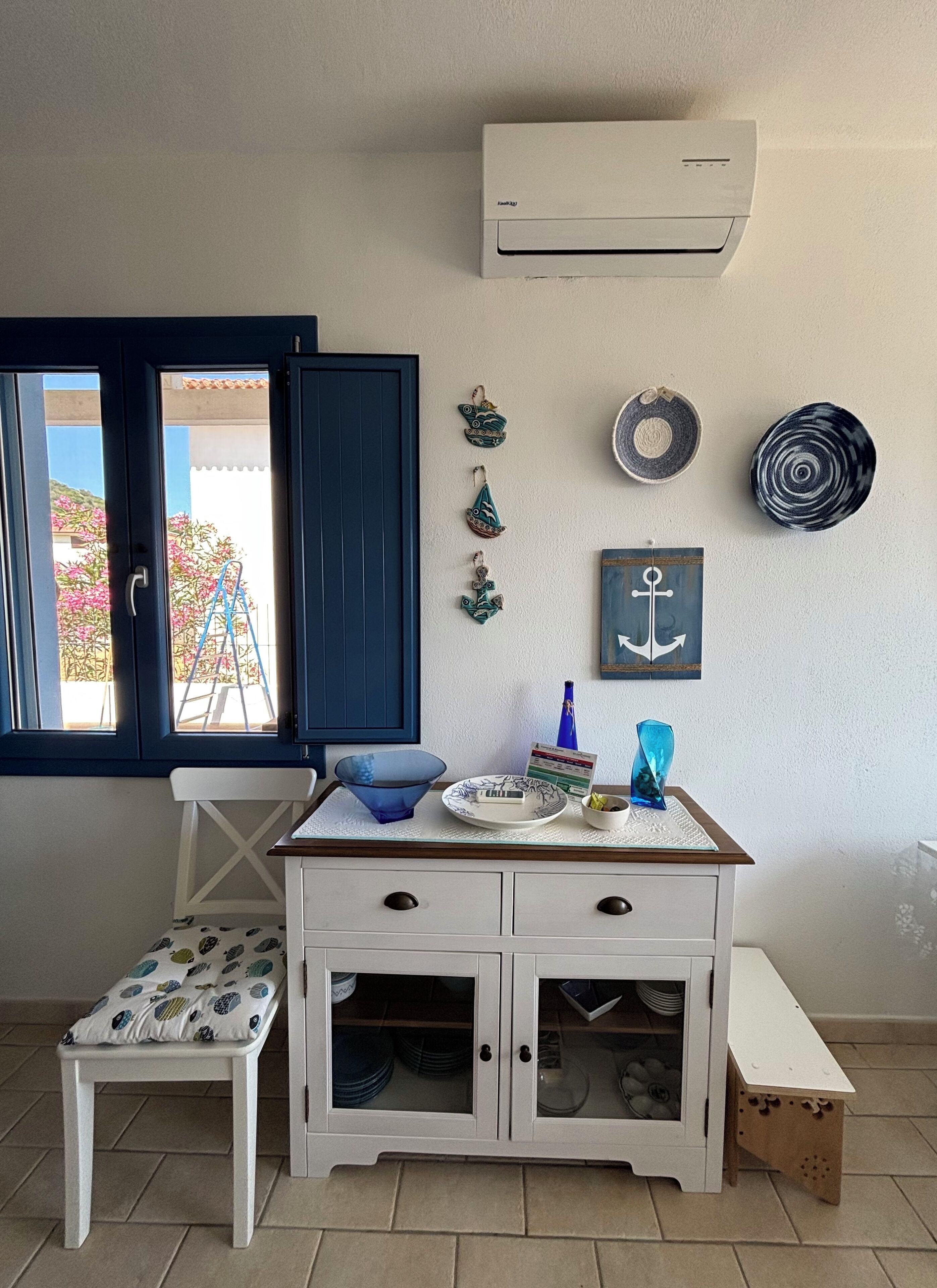 Een kamer met een witte kast, een blauwe stoel, een klein tafeltje met decoratieve objecten, een raam met uitzicht op roze bloemen en een airconditioning op de muur.