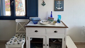Une pièce avec un meuble blanc, une chaise bleue, une petite table ornée d’objets décoratifs, une fenêtre donnant sur des fleurs roses et un climatiseur fixé au mur.