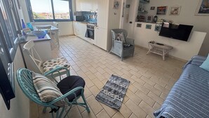 Un espace de vie compact comprenant un coin cuisine, un coin repas, un coin salon avec une chaise et une petite table, et un téléviseur fixé au mur.