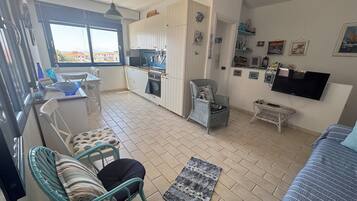 Een knus woongedeelte met een kitchenette, een eethoek, een zithoek met een stoel en een kleine tafel, en een aan de muur bevestigde televisie.