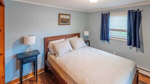 Un dormitorio con cama, mesita de noche, lámpara, ventana con cortinas y un cuadro en la pared.