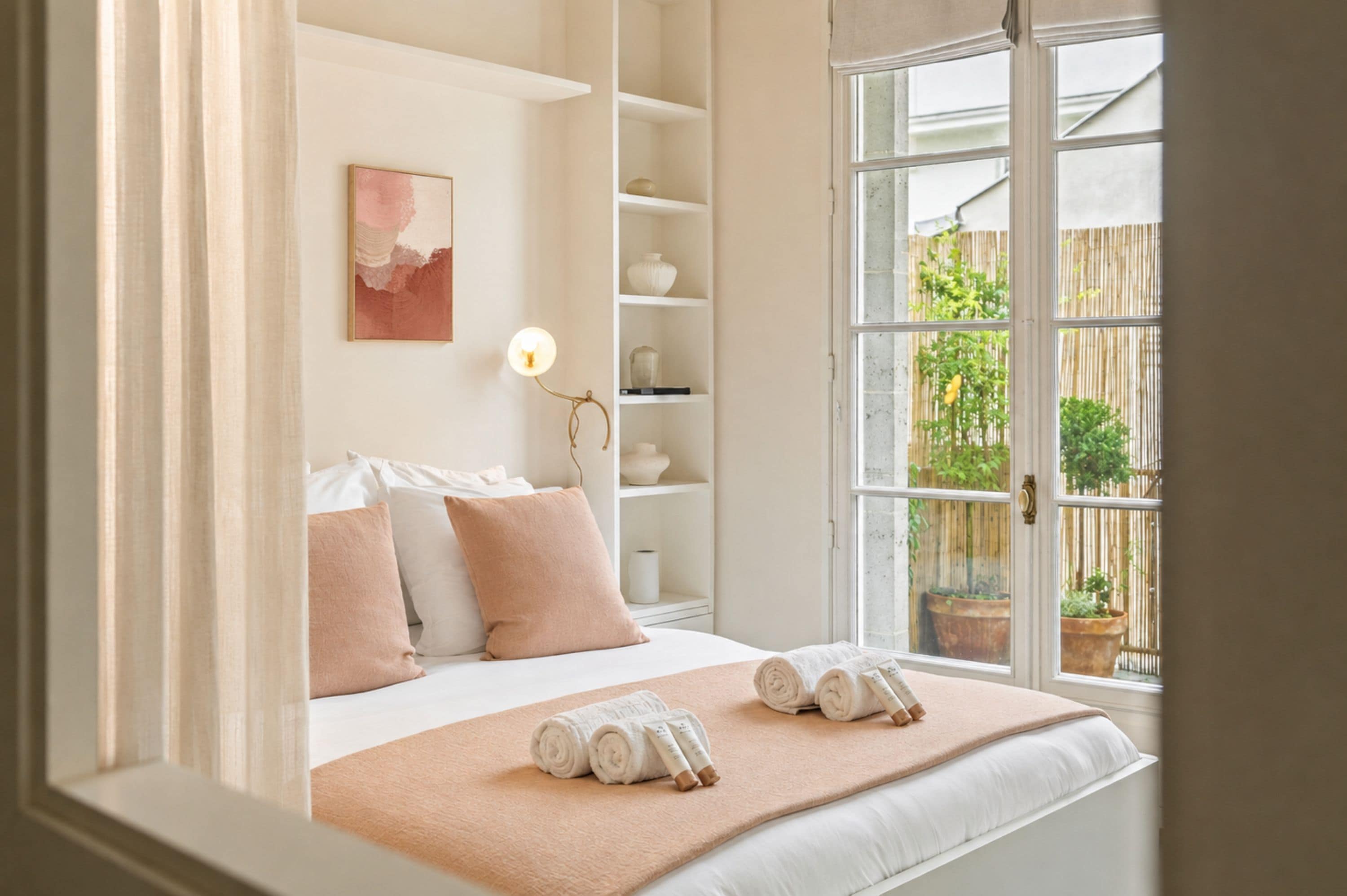 Une chambre d’hôtel comprenant un lit, des oreillers, une lampe de chevet, une fenêtre donnant sur l’extérieur et une étagère avec des objets décoratifs.