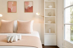 Une chambre à coucher bien rangée, avec un lit, des oreillers, une table de chevet, une lampe et une étagère ornée d’objets décoratifs.