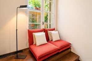 Une pièce avec un canapé rouge, une lampe sur pied et une fenêtre ornée de plantes.