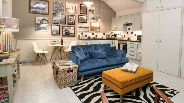 A modern living room with a blue sofa, a yellow ottoman, and a zebra-patterned rug. There is a dining area with a white table and chairs, and a kitchen with white cabinets. - Cosy Cottage (Dereham)