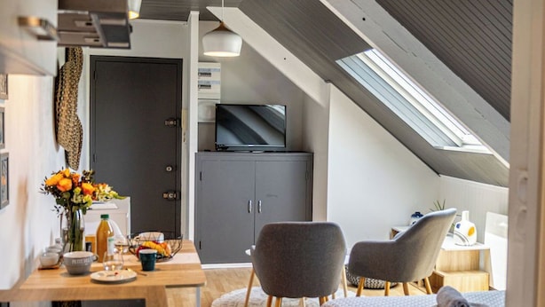 A modern loft bedroom with a sloped ceiling, a large bed, two chairs, a desk, and a television.