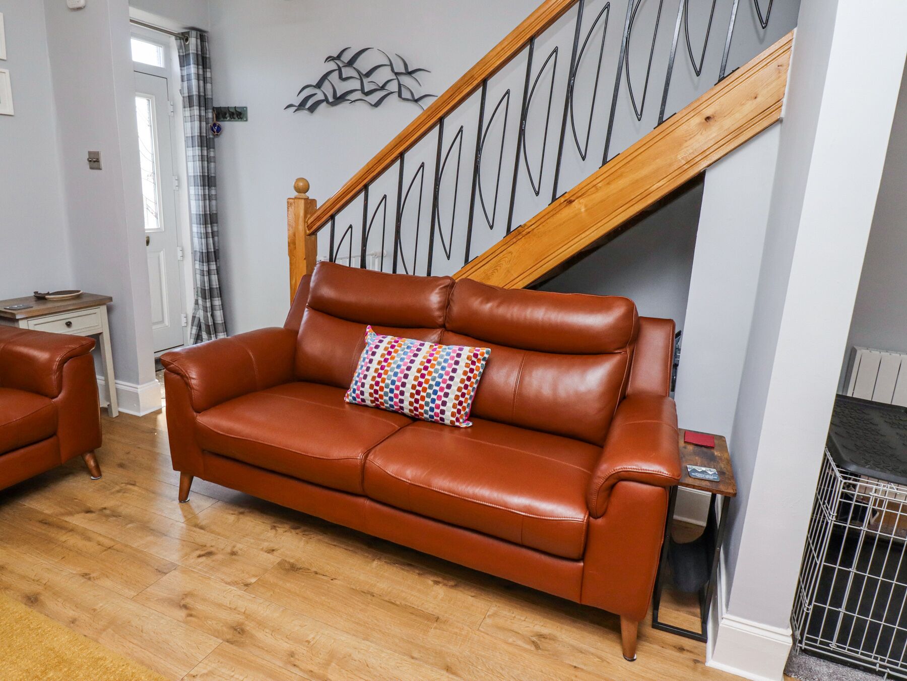 Un sofá de cuero marrón con un cojín colorido en una habitación con piso de madera, una escalera con barandilla de madera y una reja de metal.