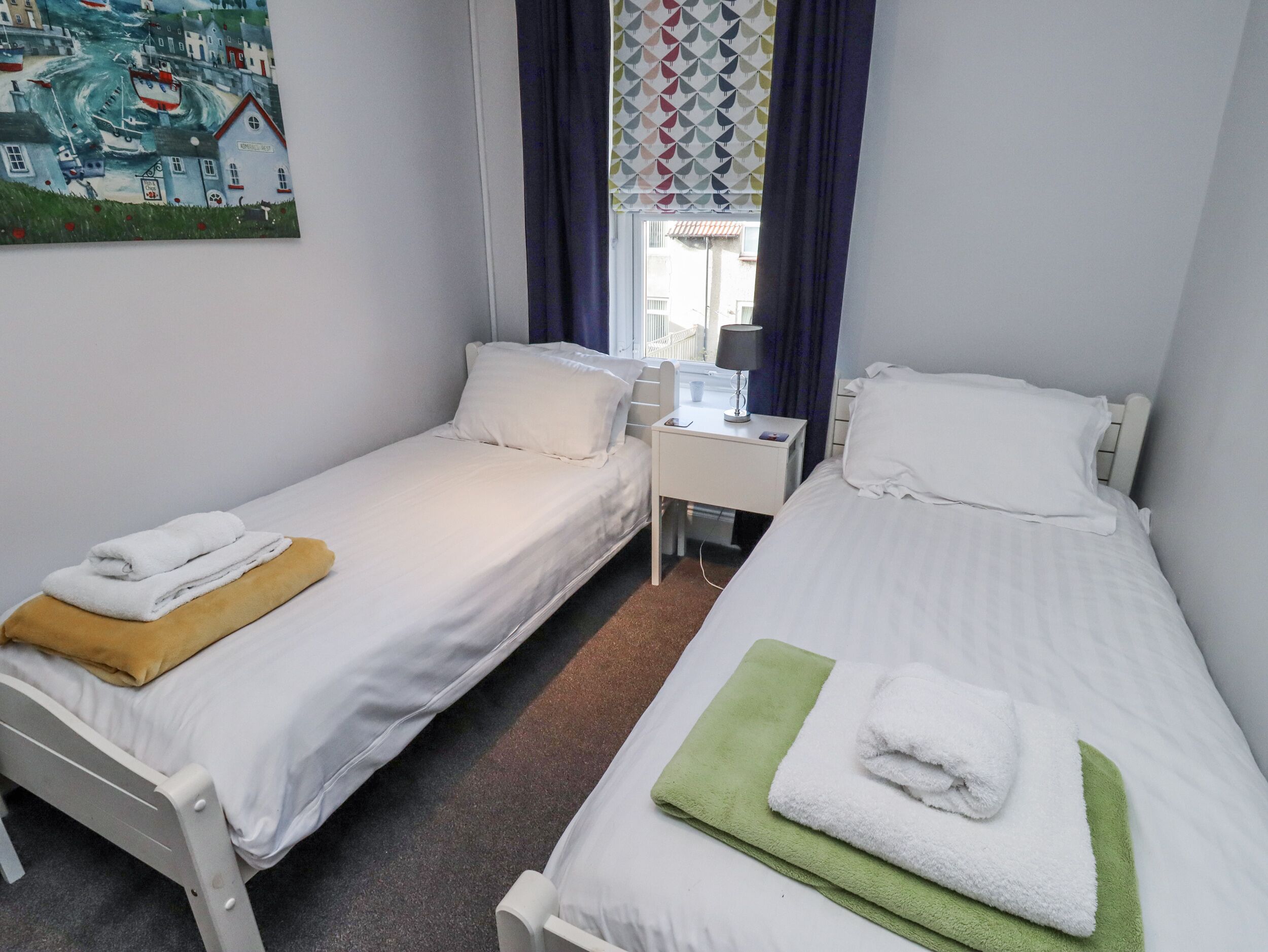 A hotel room with two single beds, a small bedside table with a lamp, a window with curtains, and a painting on the wall.