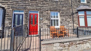 Un edificio de piedra con puerta roja, dos ventanas con contraventanas y un pequeño espacio al aire libre con sillas de madera.