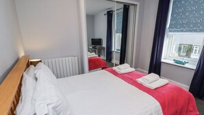 Una cama doble con ropa de cama blanca y roja, un cabecero de madera, un radiador, un espejo y una ventana con cortinas.