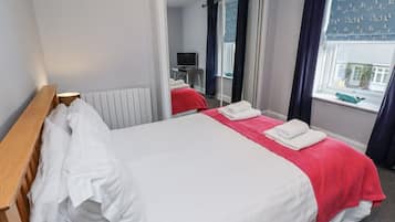 A double bed with white and red linens, a wooden headboard, a radiator, a mirror, and a window with curtains.