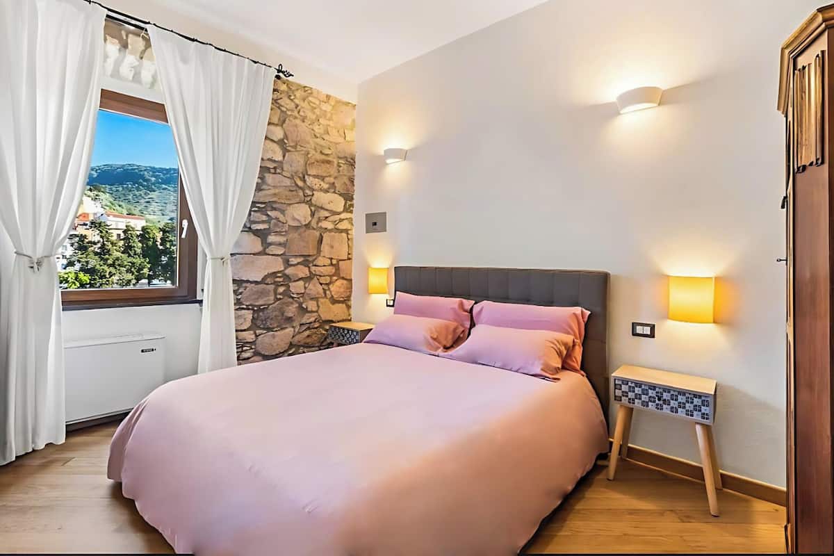 A bedroom with a stone wall, a large bed with pink bedding, two bedside lamps, and a view of greenery through a window with white curtains.