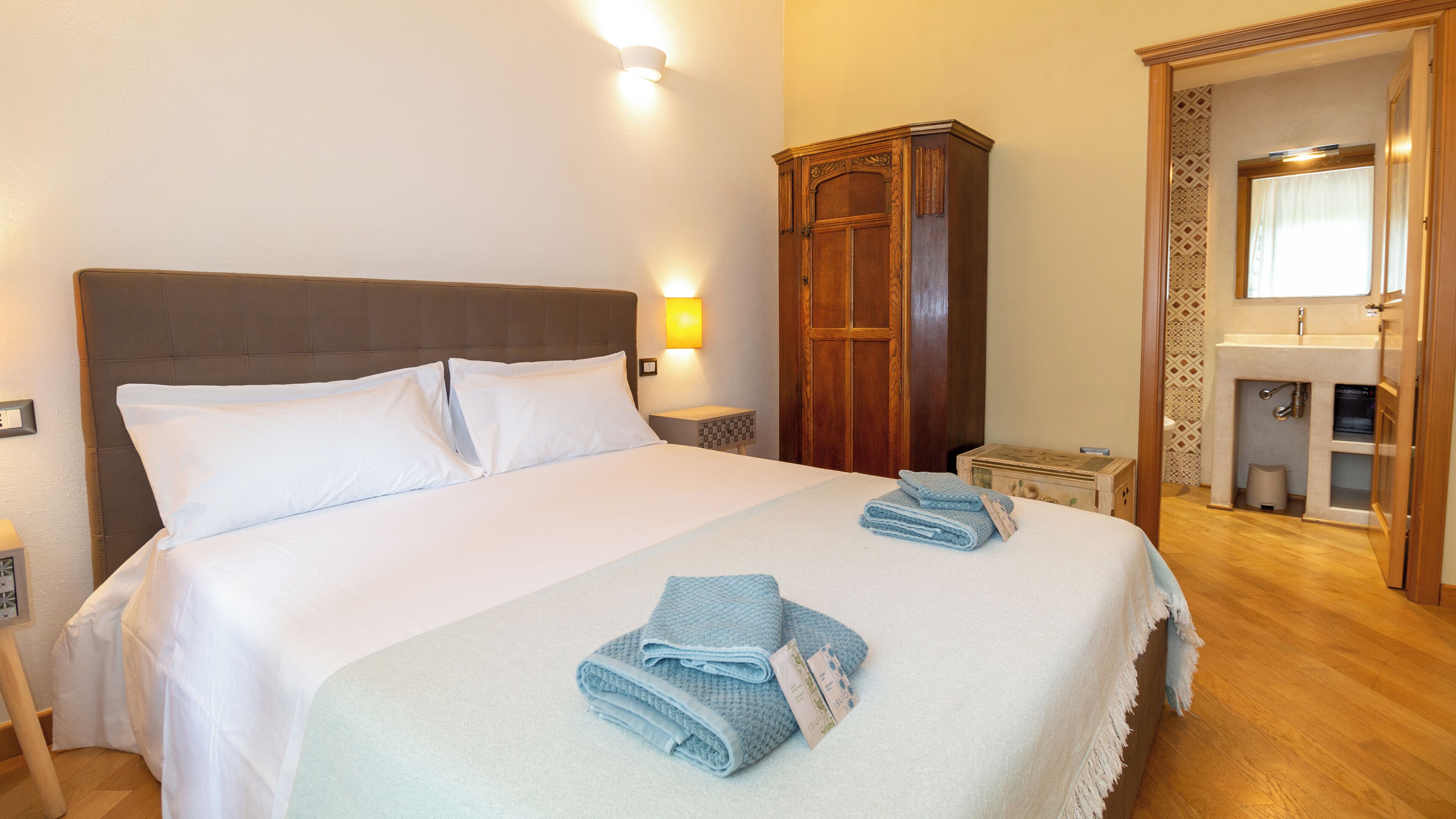 Een net opgemaakt bed met witte lakens, een houten garderobe en een badkamer met een wastafel en spiegel.