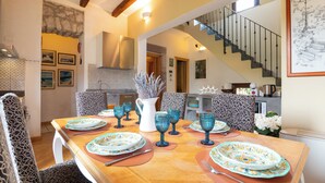 A dining area with a wooden table set for four, surrounded by patterned chairs. A staircase is visible in the background leading to an upper level. - Bosa Eye House 1873 (Bosa)