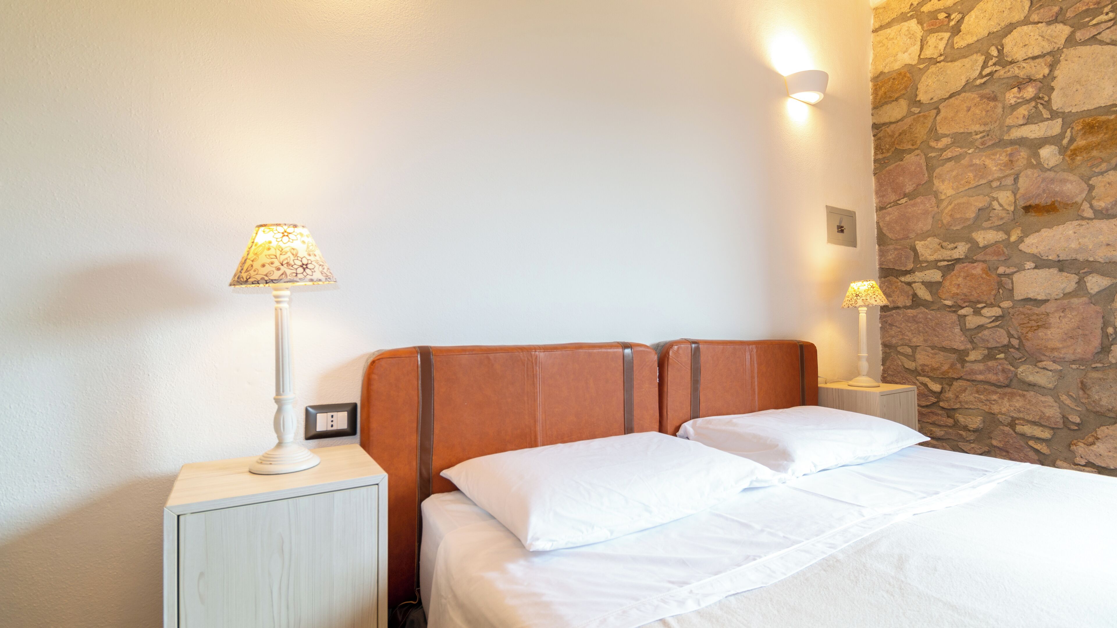 A hotel room with a bed, two bedside lamps, a nightstand, and a stone wall.