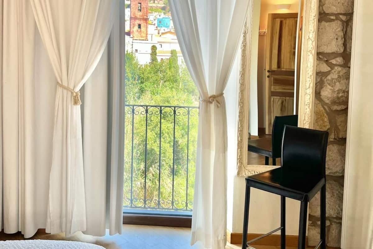 A bedroom with a balcony, white curtains, a bed, and a chair.
