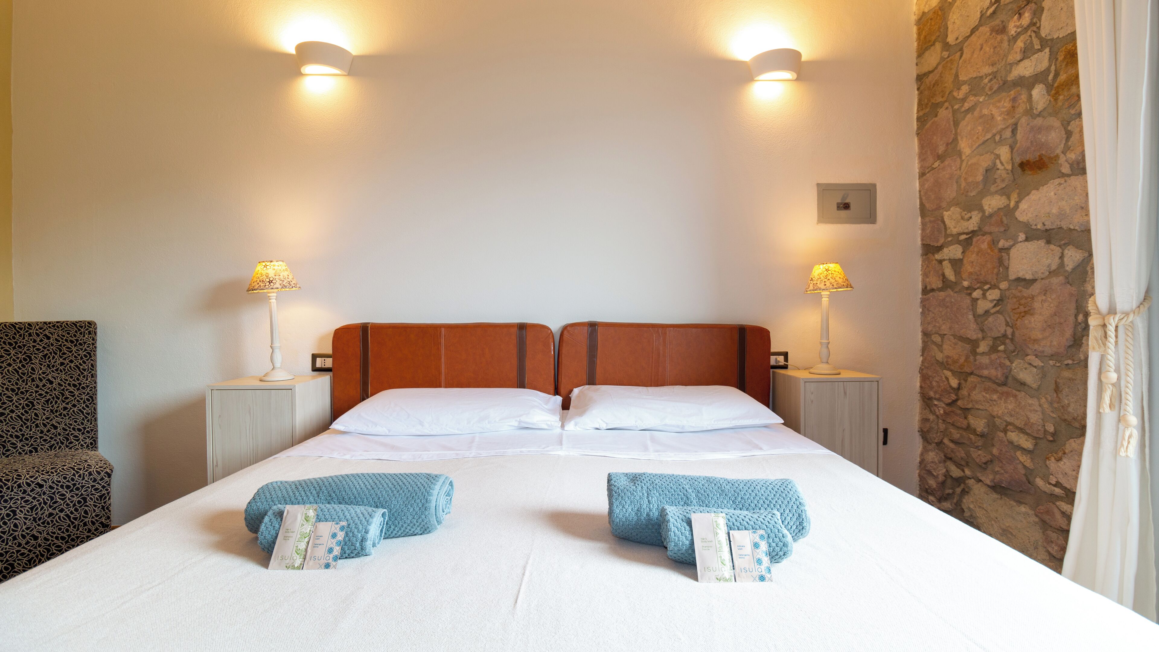 A double bed with two pillows, two bedside tables with lamps, a patterned chair, and a stone wall.