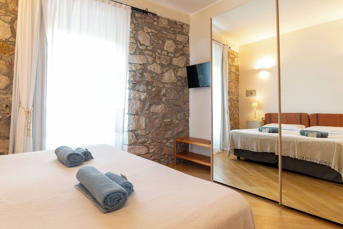 A bedroom with a stone wall, a large mirror, a bed with two pillows, and a TV mounted on the wall.