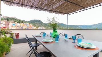 An outdoor dining area with a table set for two, surrounded by chairs, a vase with flowers, and a view of colorful buildings and hills in the background.